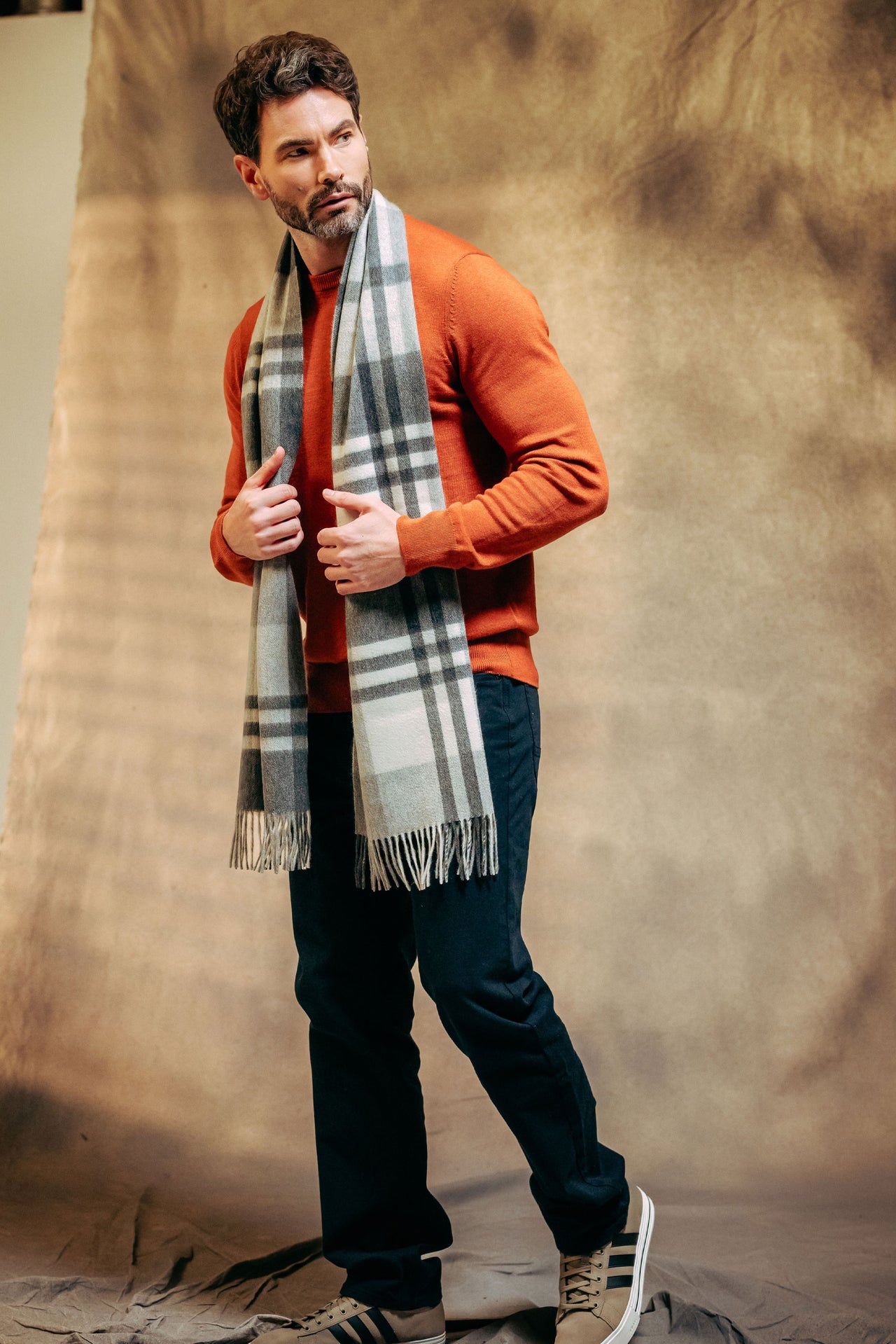 Man wearing an orange sweater, dark pants, and a plaid cashmere scarf against a textured brown background