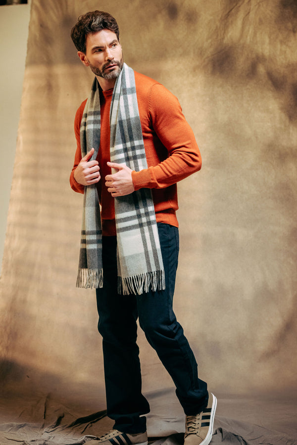 Man wearing an orange sweater, dark pants, and a plaid cashmere scarf against a textured brown background