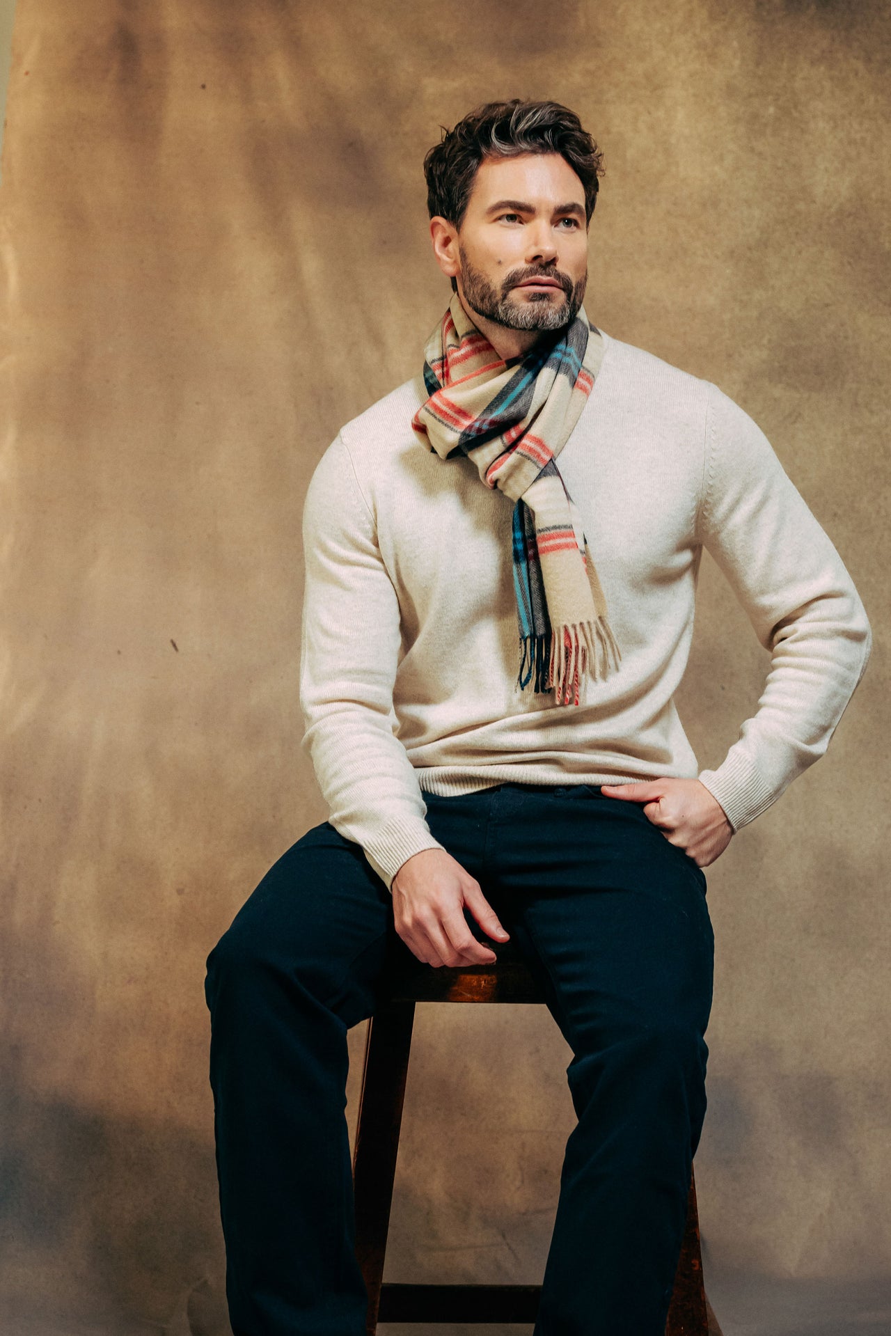 Man wearing a white sweater and plaid cashmere scarf sitting on a stool against a brown background