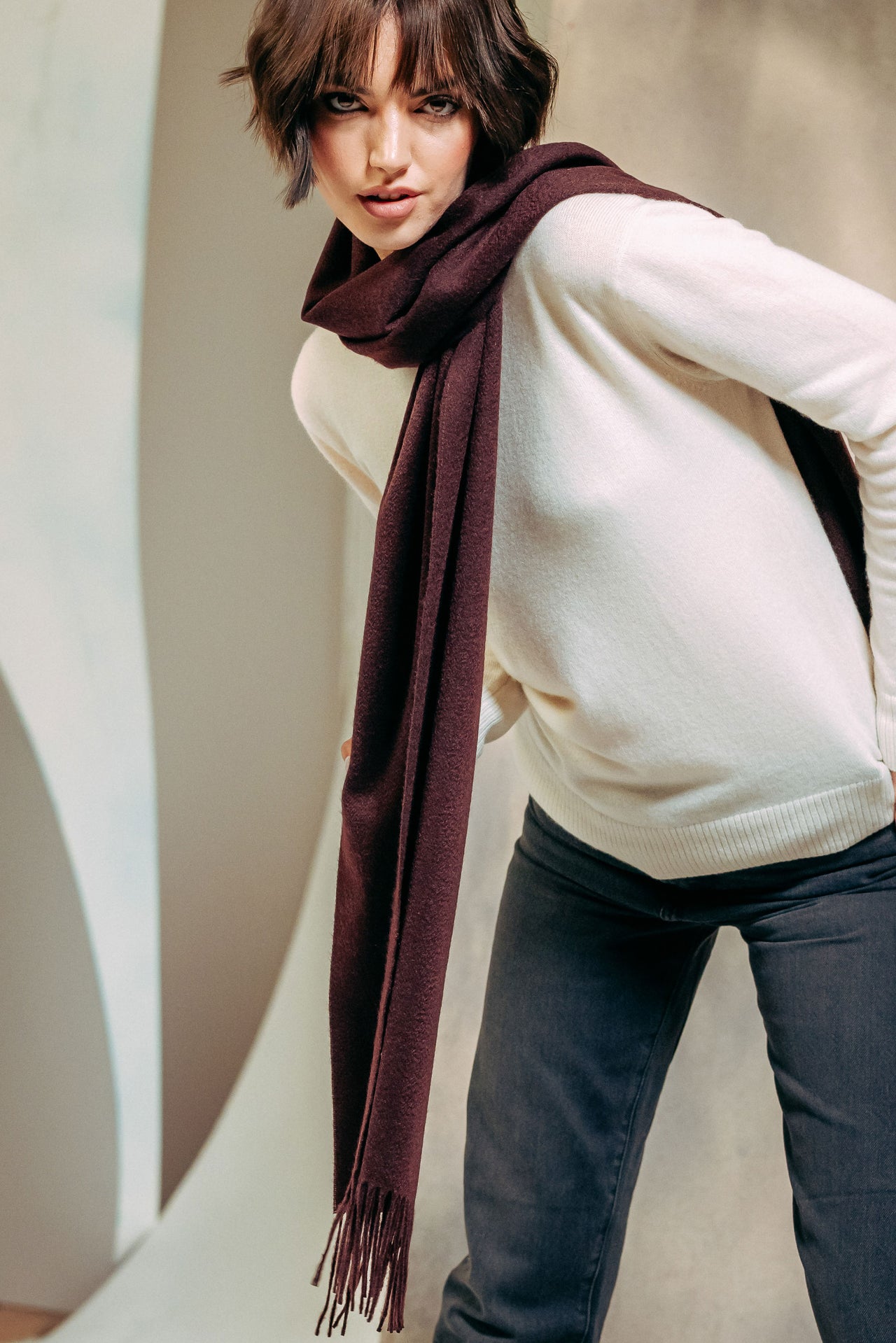 Woman wearing a white sweater and dark jeans with a burgundy cashmere scarf against a neutral background