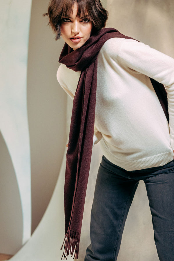 Woman wearing a white sweater and dark jeans with a burgundy cashmere scarf against a neutral background