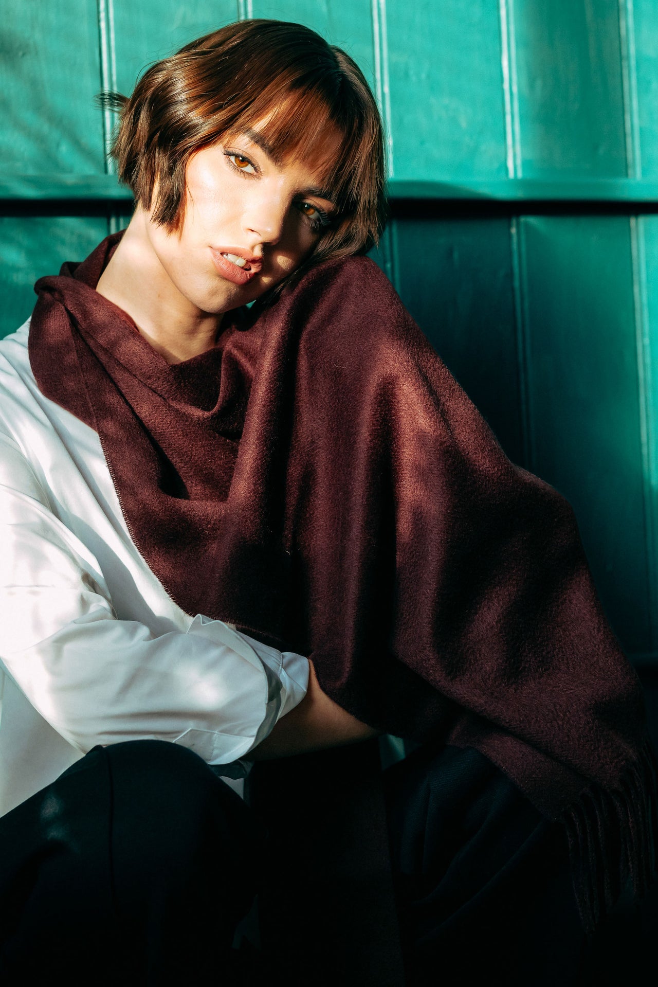 Person wearing a dark brown cashmere scarf over a white shirt against a teal tiled wall.