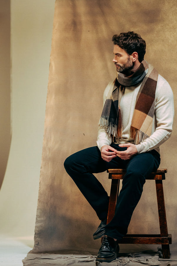 Man sitting on a wooden stool wearing a cashmere scarf and dark pants against a textured wall.