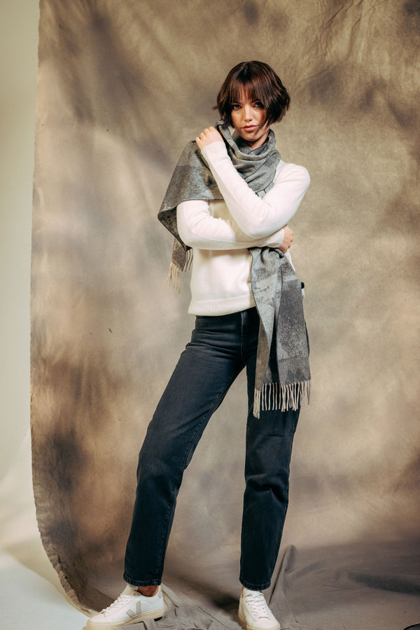 Person wearing a white sweater, black pants, and a gray plaid cashmere scarf against a textured brown background